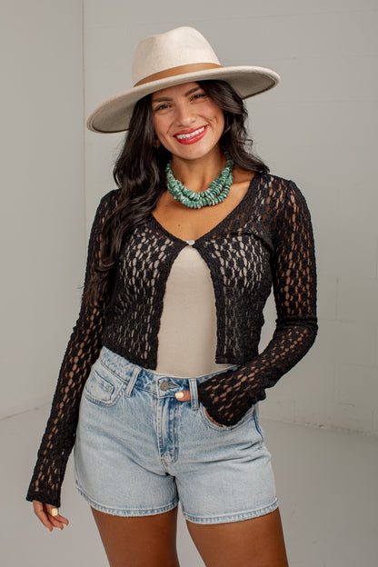 A woman modeling a black long-sleeve lace cardigan over a beige top, paired with light-wash denim shorts and a beige wide-brim hat, against a plain light gray studio background.