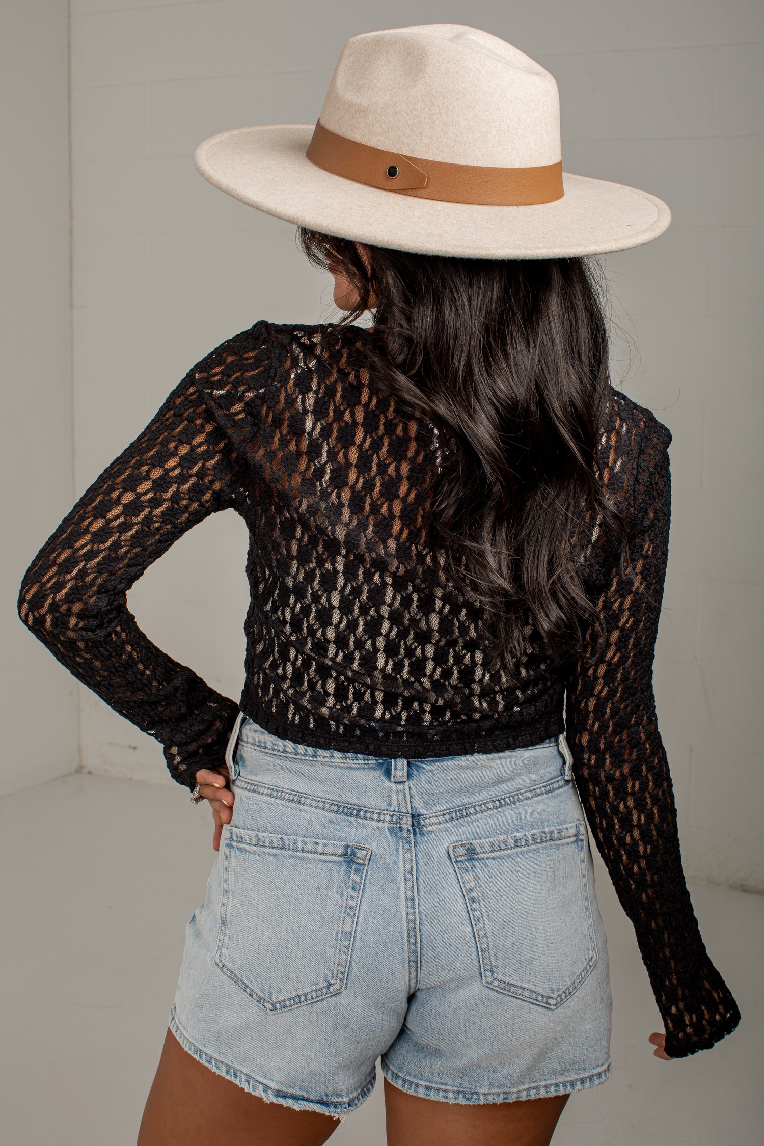 A woman shown from the back wearing a black long-sleeve lace mesh crop top, light-wash denim shorts, and a cream fedora hat with a brown band, standing against a light gray wall.