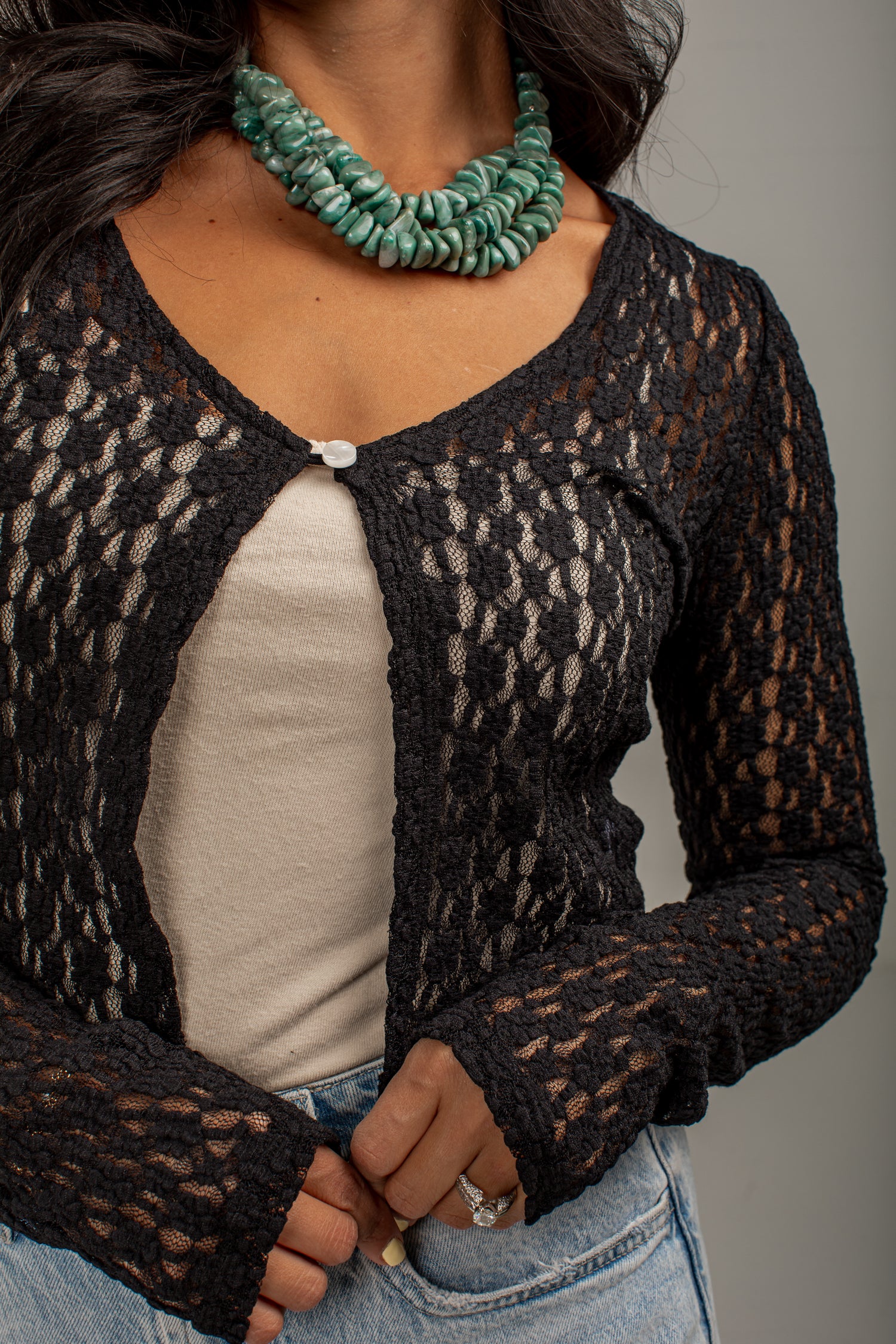 A close-up of a person wearing a black open-knit cardigan over a light beige top, paired with blue jeans and a chunky green beaded necklace, against a plain gray background.