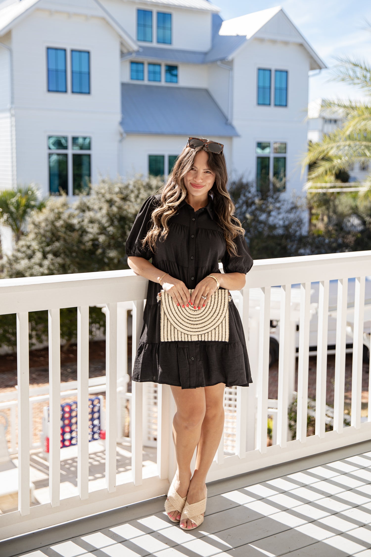 Woman in a black dress holding a woven handbag on a balcony with a house in the background