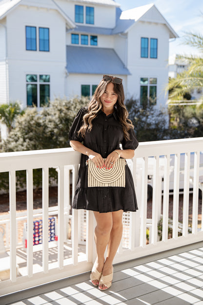 Woman in a black dress holding a woven handbag on a balcony with a house in the background
