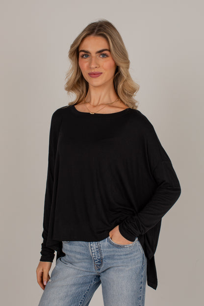 A woman modeling a black long-sleeve boat-neck top with a draped, slightly asymmetrical hem, paired with light-wash blue jeans, against a solid light gray studio background.