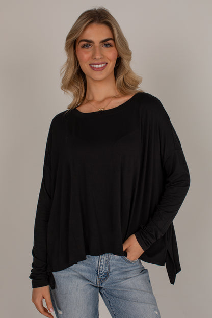 A woman with blonde hair wearing a black long-sleeve boat-neck top and light-wash blue jeans, standing against a solid light gray background.