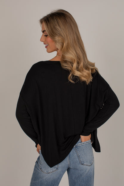 A woman with blonde hair is shown from the back wearing a loose-fitting black long-sleeve top and light blue jeans against a plain light gray studio background.