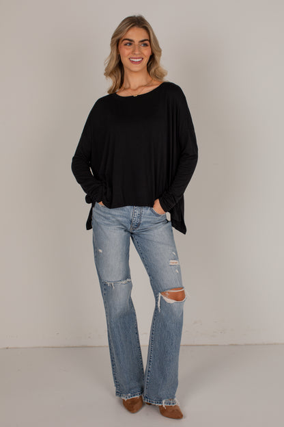 A woman modeling a black long-sleeve draped top and light-wash distressed jeans against a plain light gray studio background.