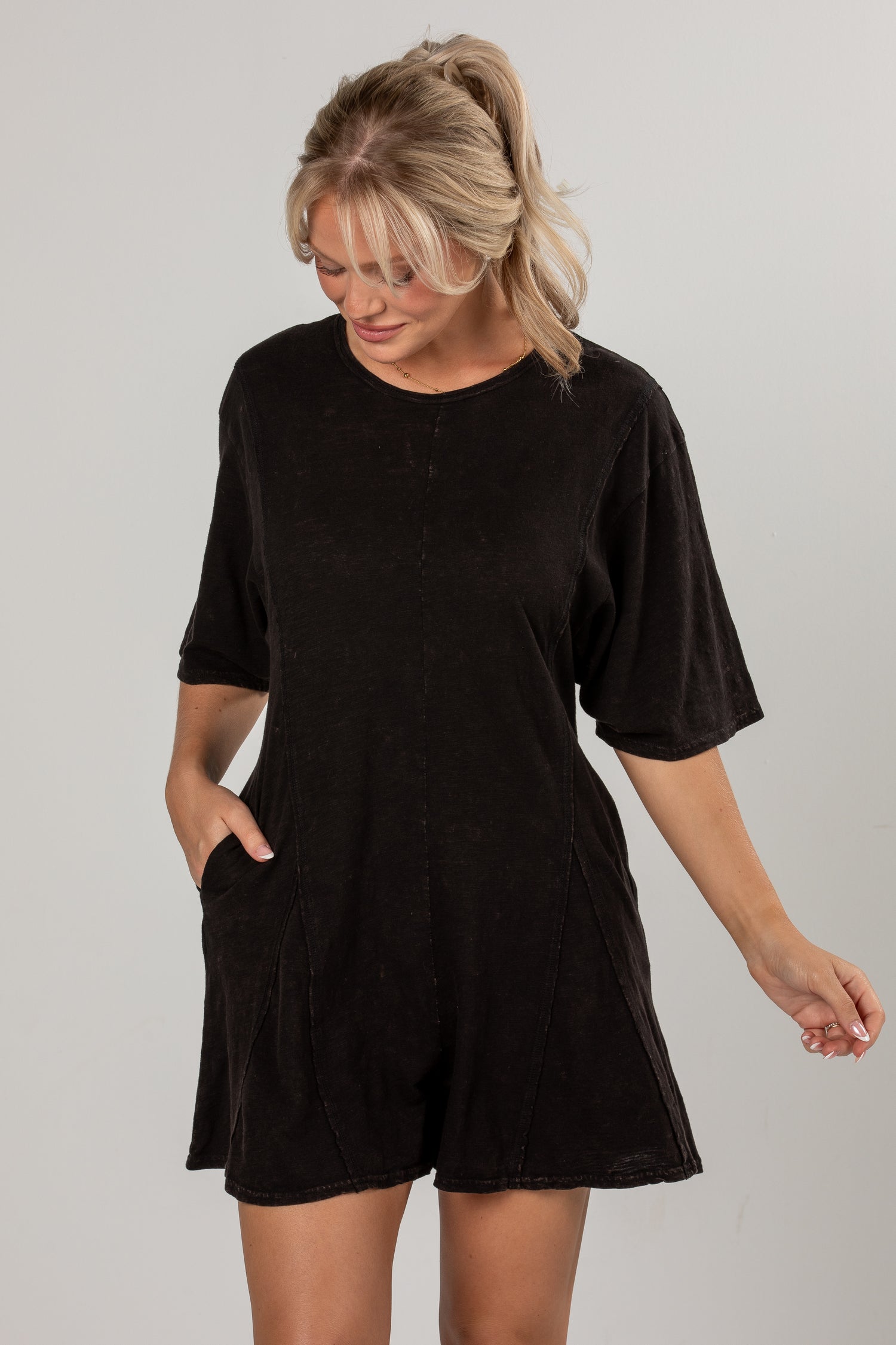 A woman wearing a black short-sleeve romper with side pockets, standing against a light gray studio background.