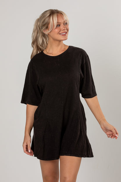 A woman modeling a short-sleeve, oversized black romper with a relaxed, slightly faded texture against a plain light gray studio background.