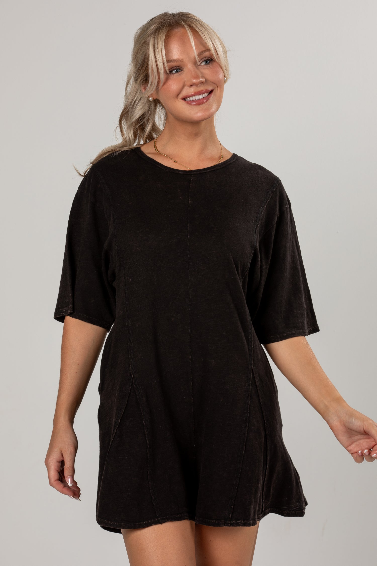 A woman modeling a short-sleeve, oversized black romper with a faded, washed texture against a plain light gray studio background.