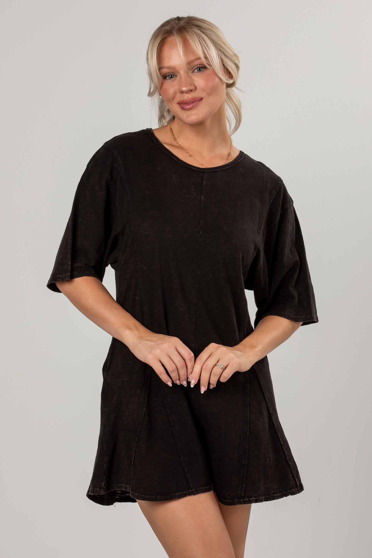 A woman modeling a black short-sleeve romper with a flared skirt, photographed against a plain light gray studio background.