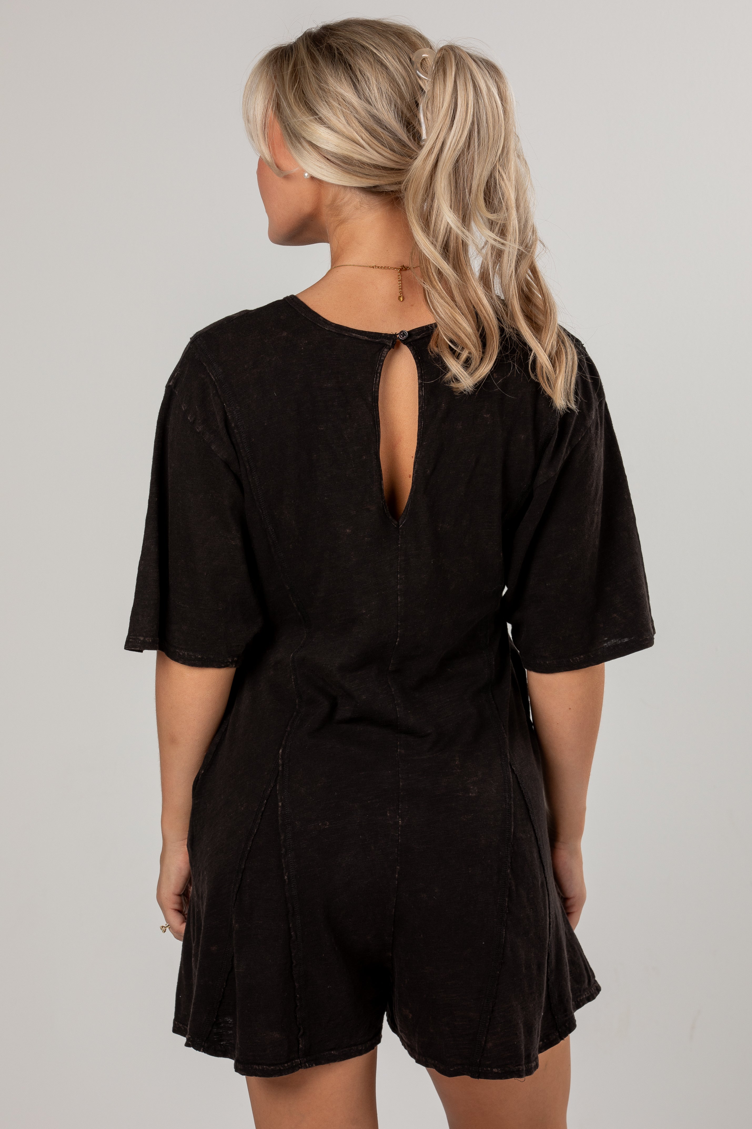 A woman shown from the back wearing a black short-sleeve romper with a keyhole cutout at the neckline, photographed against a plain light gray studio background.