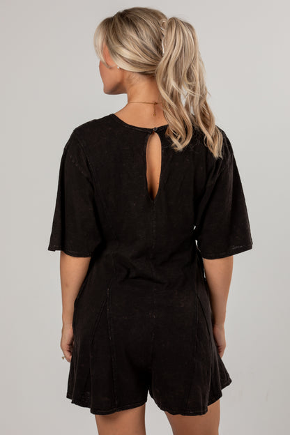 A woman shown from the back wearing a black short-sleeve romper with a keyhole cutout at the neckline, photographed against a plain light gray studio background.