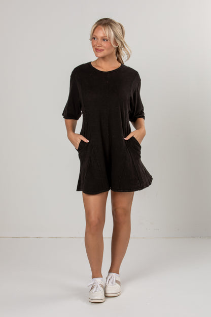A woman modeling a black short-sleeve romper with side pockets, paired with white sneakers, standing against a plain light gray studio background.