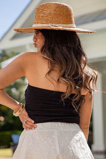 Woman wearing a straw hat, black top, and white pants with a blurred outdoor background