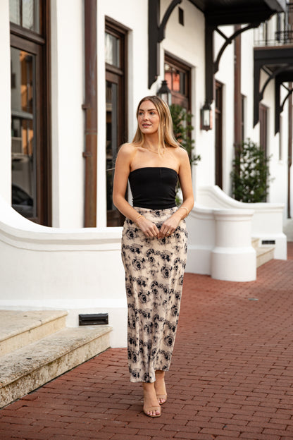 Woman wearing a black strapless top and floral skirt standing on a brick sidewalk.