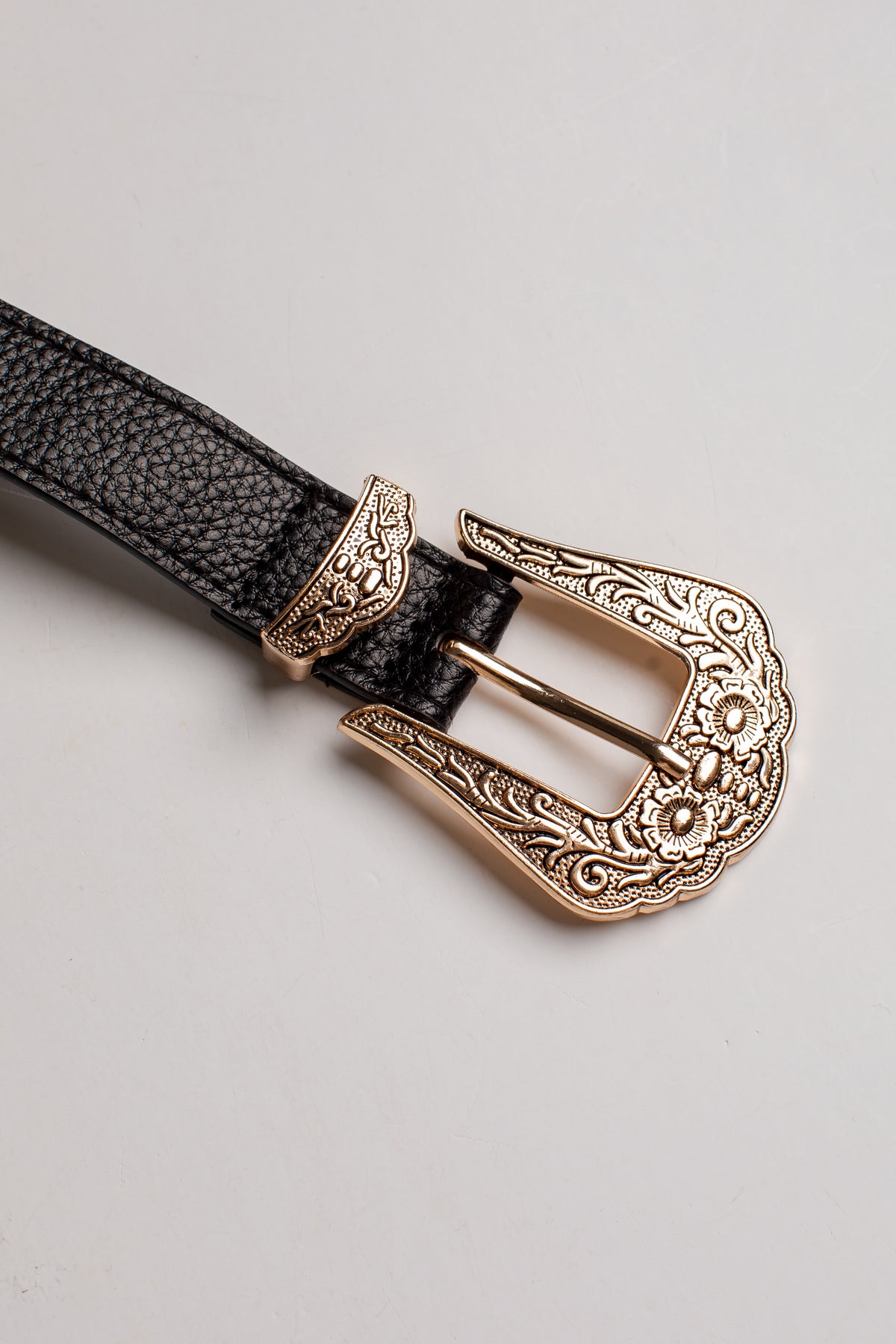 A close-up of a black textured leather belt with an ornate, engraved rose gold metal buckle, set against a plain light gray background.