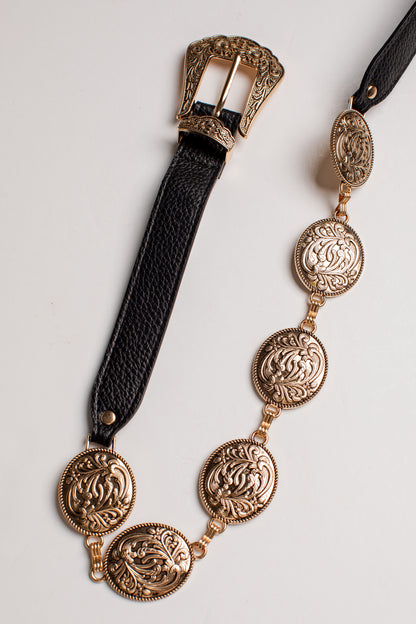 A black leather belt with ornate gold-toned metal hardware, including a large engraved buckle and a series of matching oval medallion links along the strap, displayed on a light neutral background.