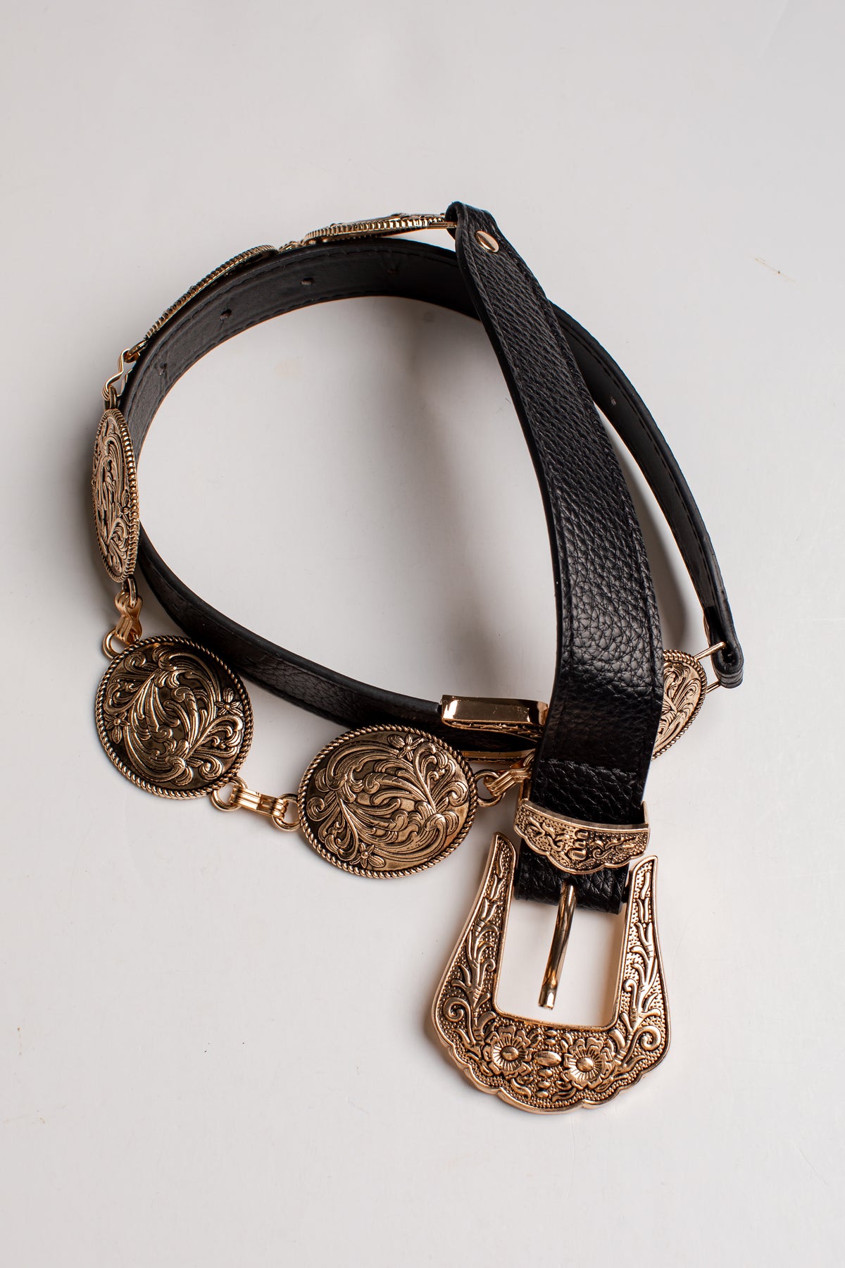 A western-style belt with a textured black leather strap and ornate antique bronze-toned metal hardware, including a large engraved buckle, medallion chain, and decorative coins, displayed on a light gray background.