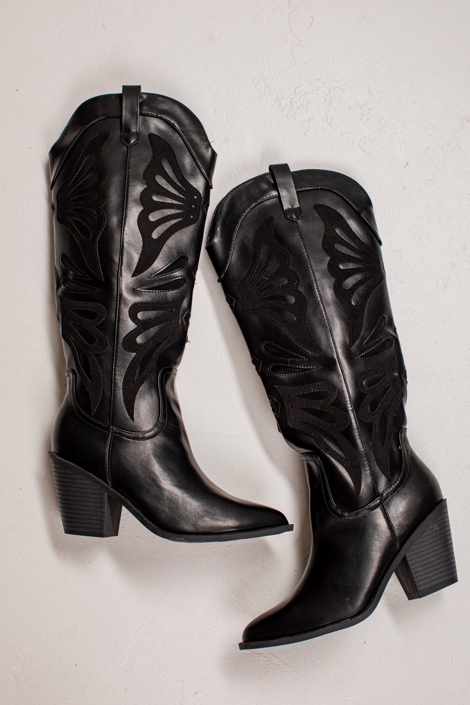 A pair of black leather knee-high cowboy boots with decorative cutout stitching, displayed on a light textured background.