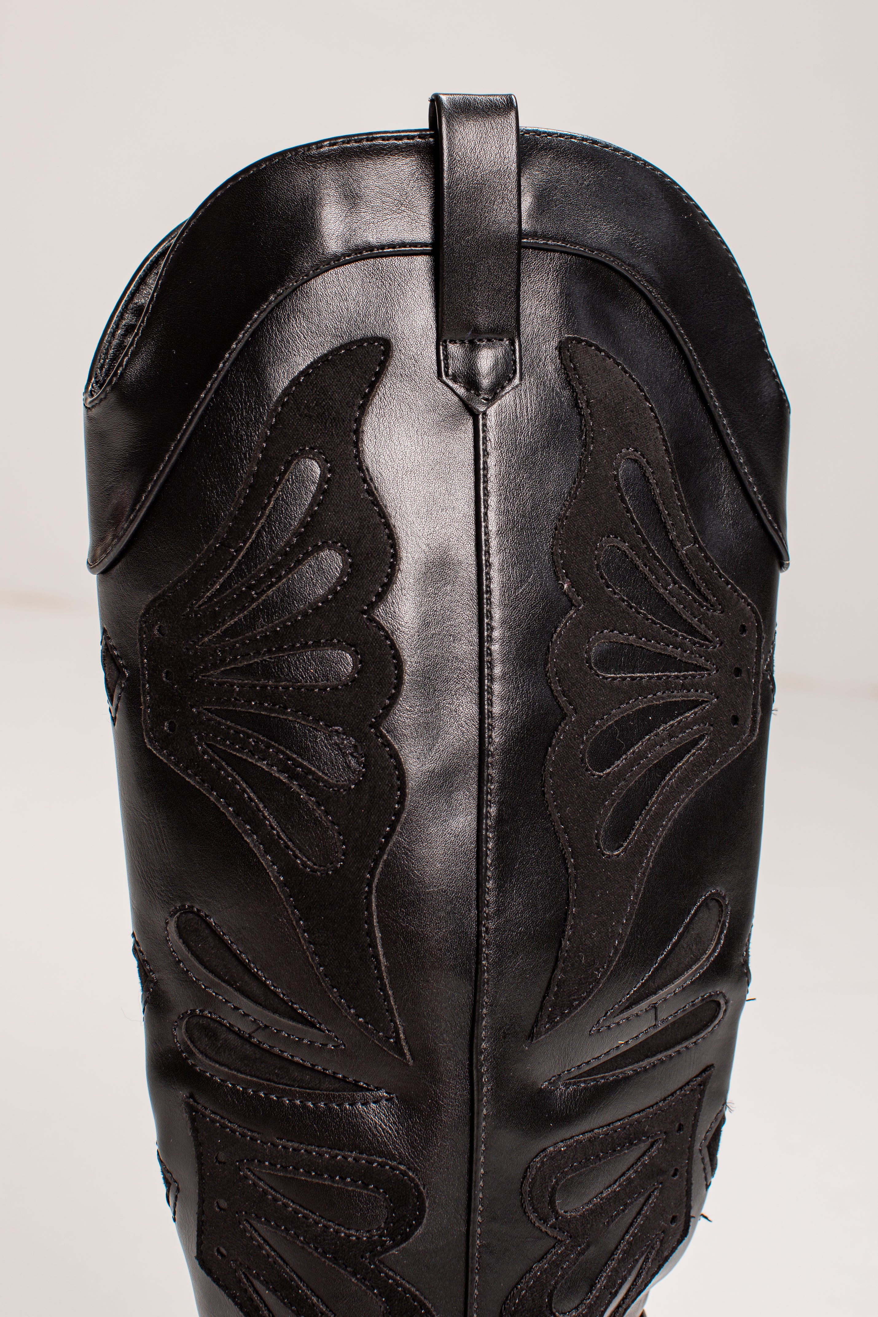 Close-up of a black leather cowboy boot shaft with decorative cutout stitching and a pull tab at the top, photographed against a plain light background.