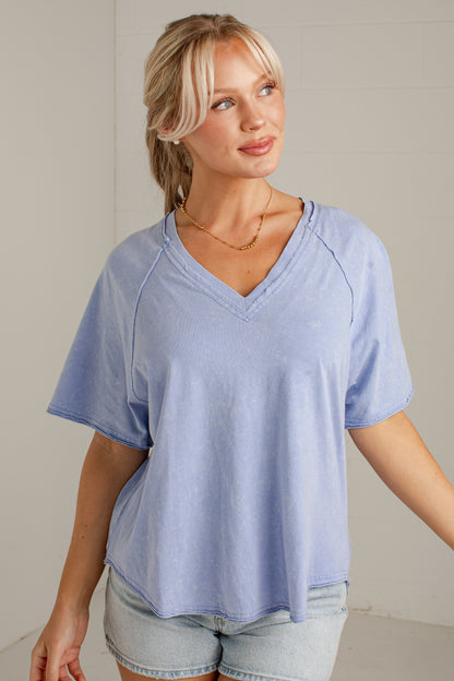 A woman wearing a light blue short-sleeve V-neck top with raglan sleeves and light denim shorts, standing against a light gray wall.