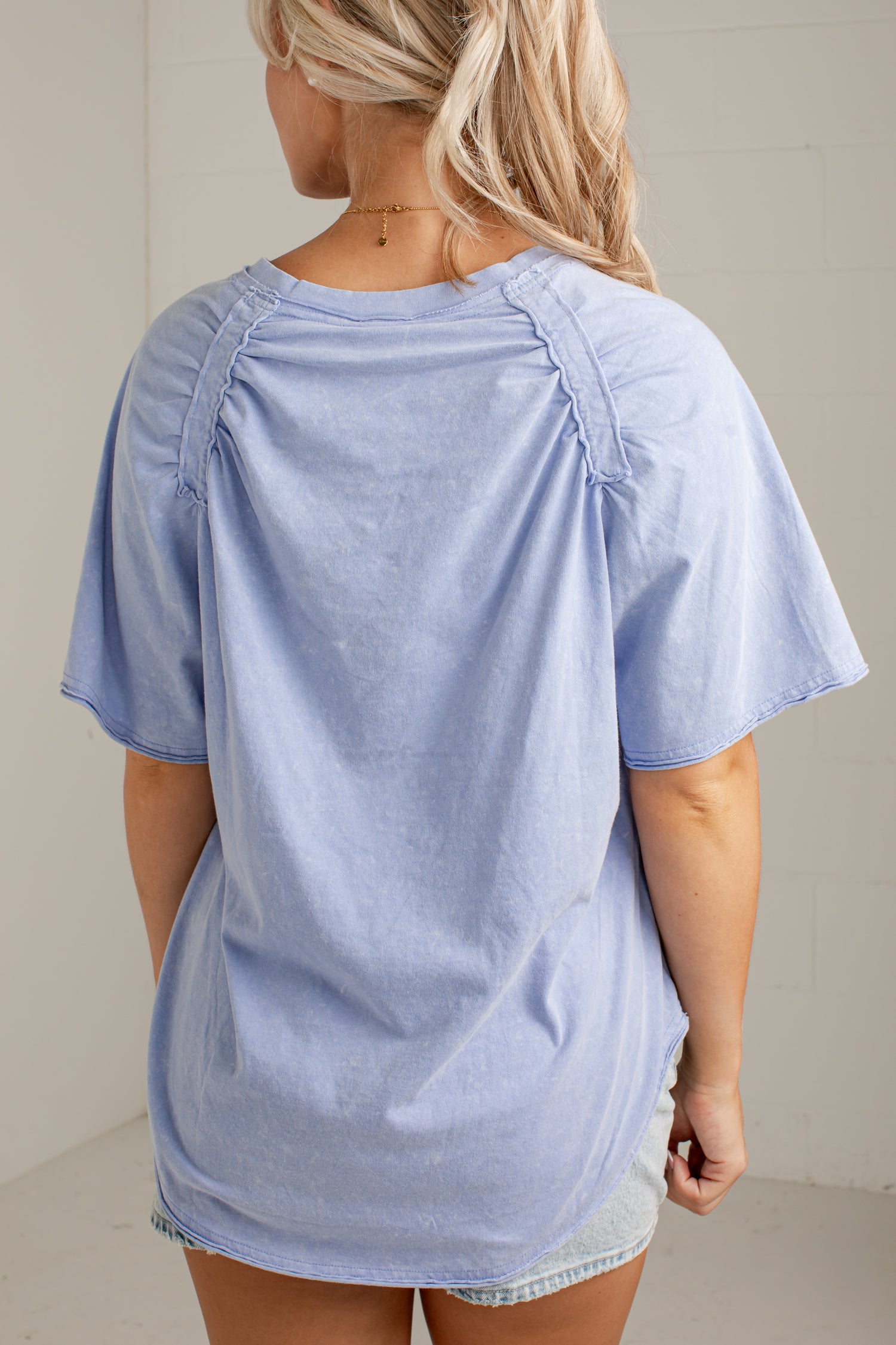 A woman viewed from the back wearing an oversized light blue short-sleeve top with gathered shoulder details and a relaxed fit, paired with light denim shorts in a simple indoor setting.