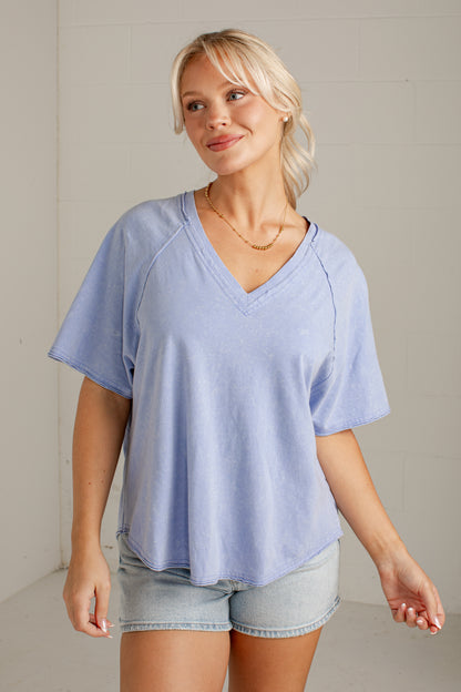 A woman wearing a light periwinkle blue V-neck short-sleeve top with raglan sleeves and light-wash denim shorts, standing against a neutral off-white wall.