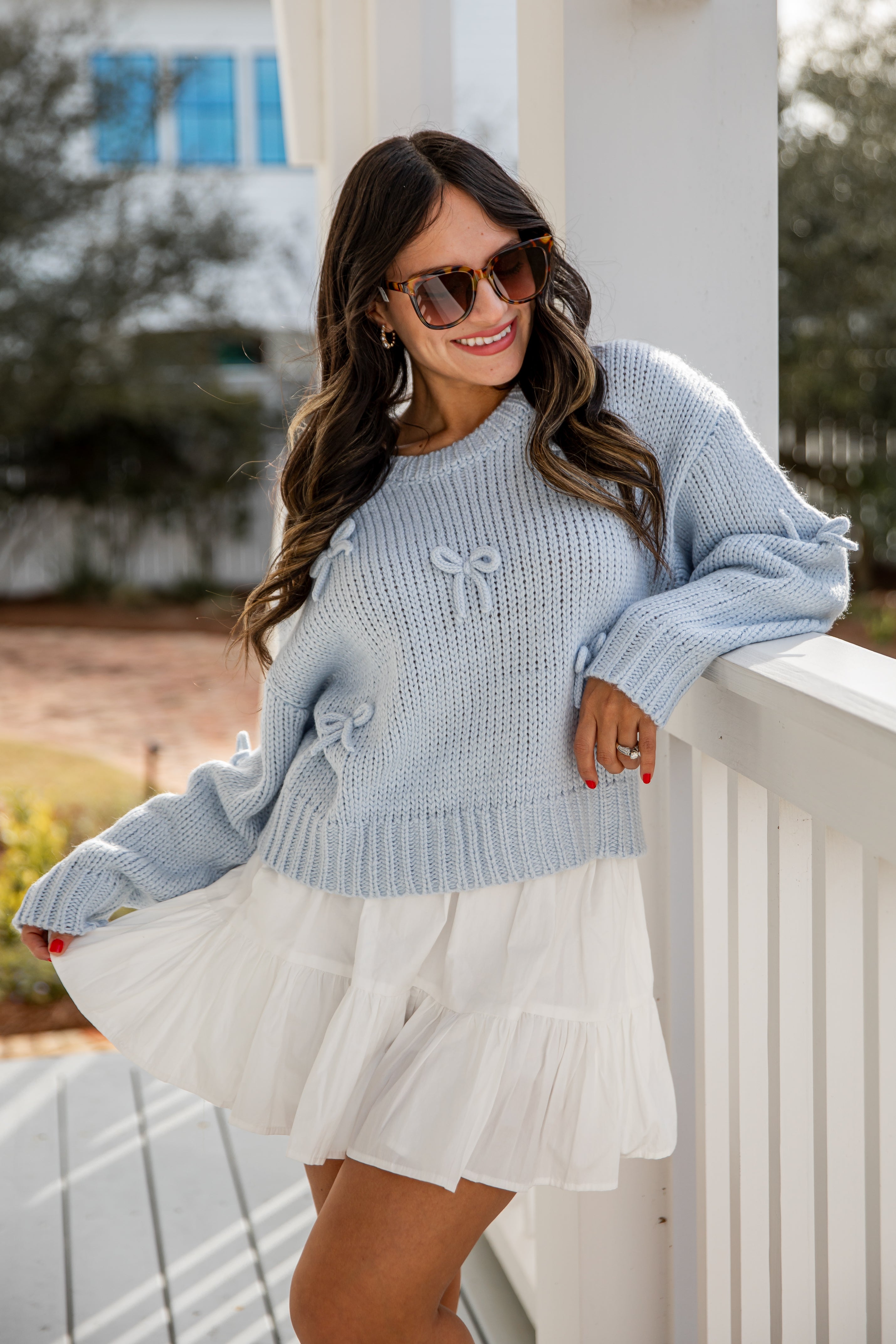 Woman wearing a light blue sweater with bow details and a white skirt, standing on a porch.