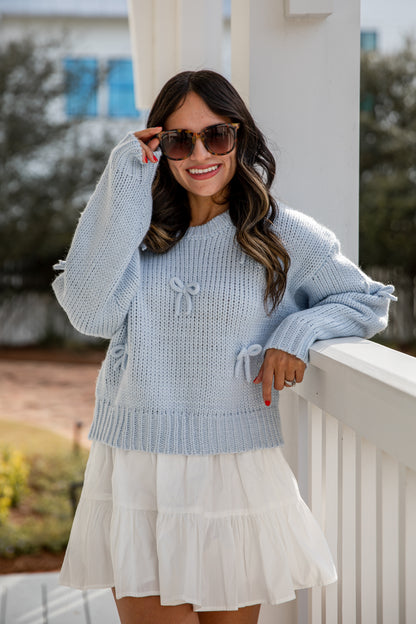 Woman wearing a light blue sweater and white skirt standing on a porch.
