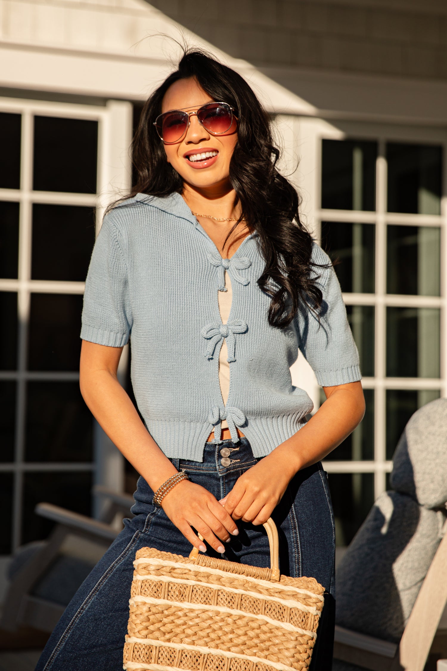 Woman wearing a light blue cardigan and sunglasses, holding a woven bag outdoors.