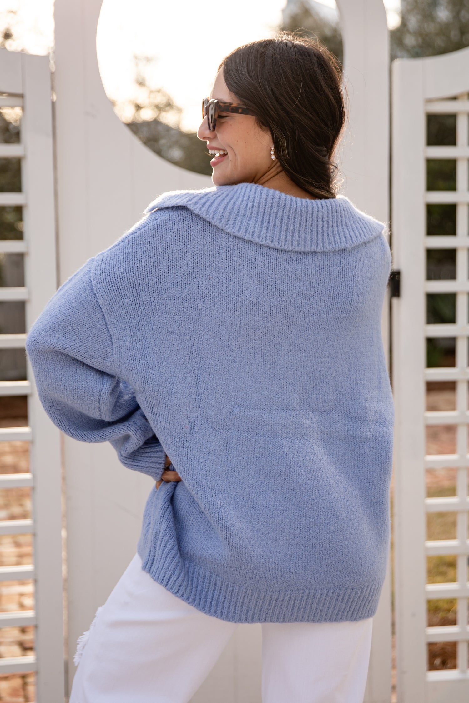 Woman wearing a light blue sweater and white pants standing in front of a white lattice fence.