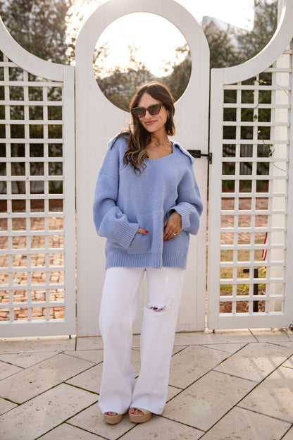 Woman wearing a blue sweater and white pants standing in front of a white lattice gate.