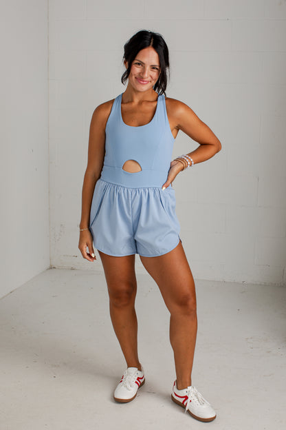 A woman modeling a light blue athletic romper with a side cutout, paired with white sneakers, standing in a white studio corner.