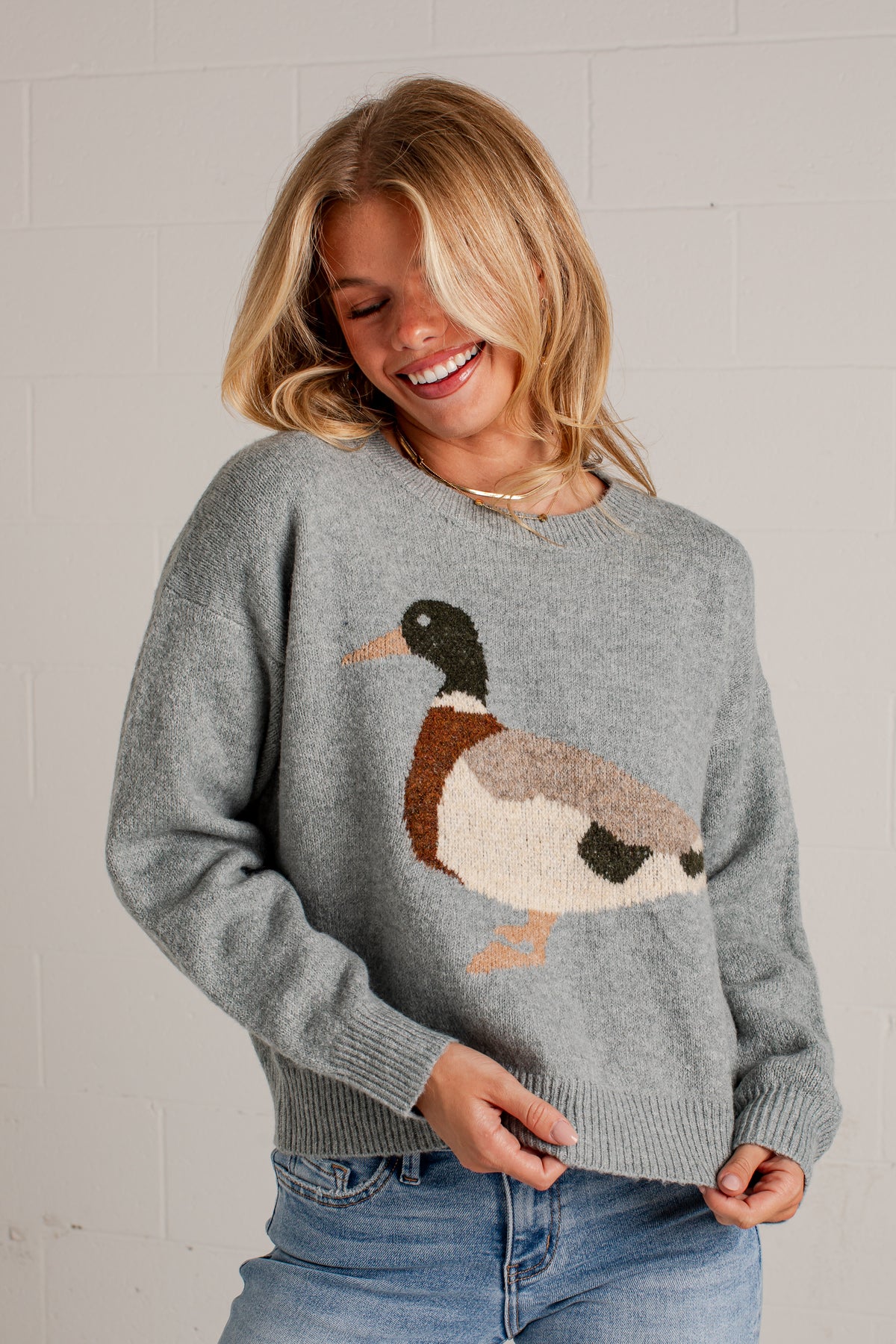 A woman smiling while wearing a blue knit sweater with a large duck motif on the front, paired with blue jeans, standing against a white brick wall.