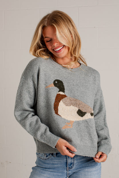 A woman smiling while wearing a blue knit sweater with a large duck motif on the front, paired with blue jeans, standing against a white brick wall.