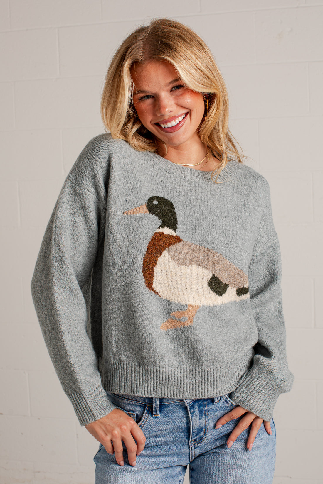 A smiling blonde woman wearing a heather gray knit sweater with a large duck motif across the chest, paired with blue jeans, standing against a white brick wall.