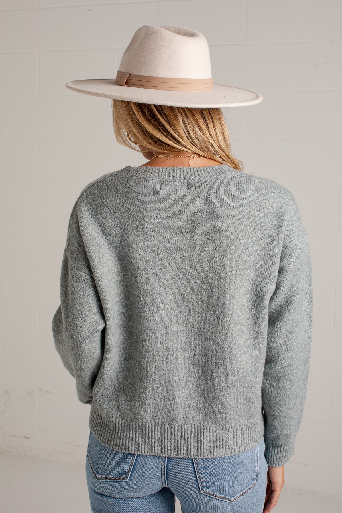 A woman shown from the back wearing a light gray knit sweater, blue jeans, and a cream-colored wide-brim hat against a white brick wall.