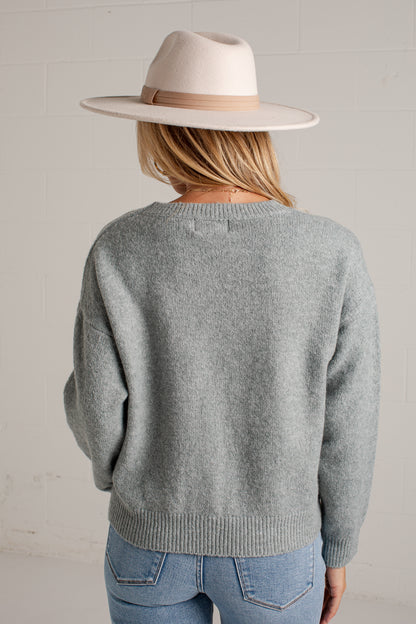 A woman shown from the back wearing a light gray knit sweater, blue jeans, and a cream-colored wide-brim hat against a white brick wall.