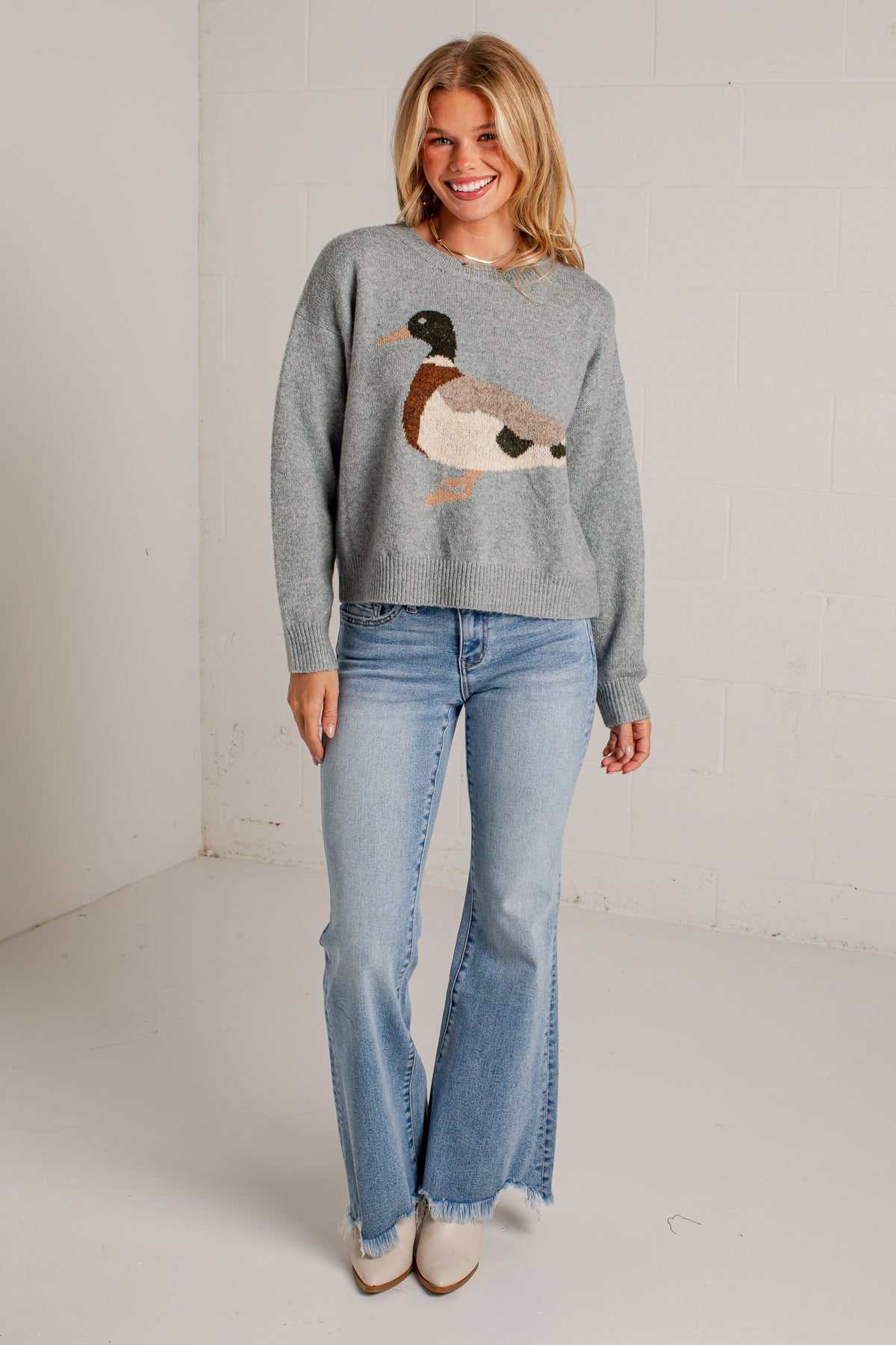 A woman modeling a heather gray crewneck sweater with a large embroidered mallard duck on the front, paired with light-wash flare jeans and beige ankle boots, standing against a white brick wall.