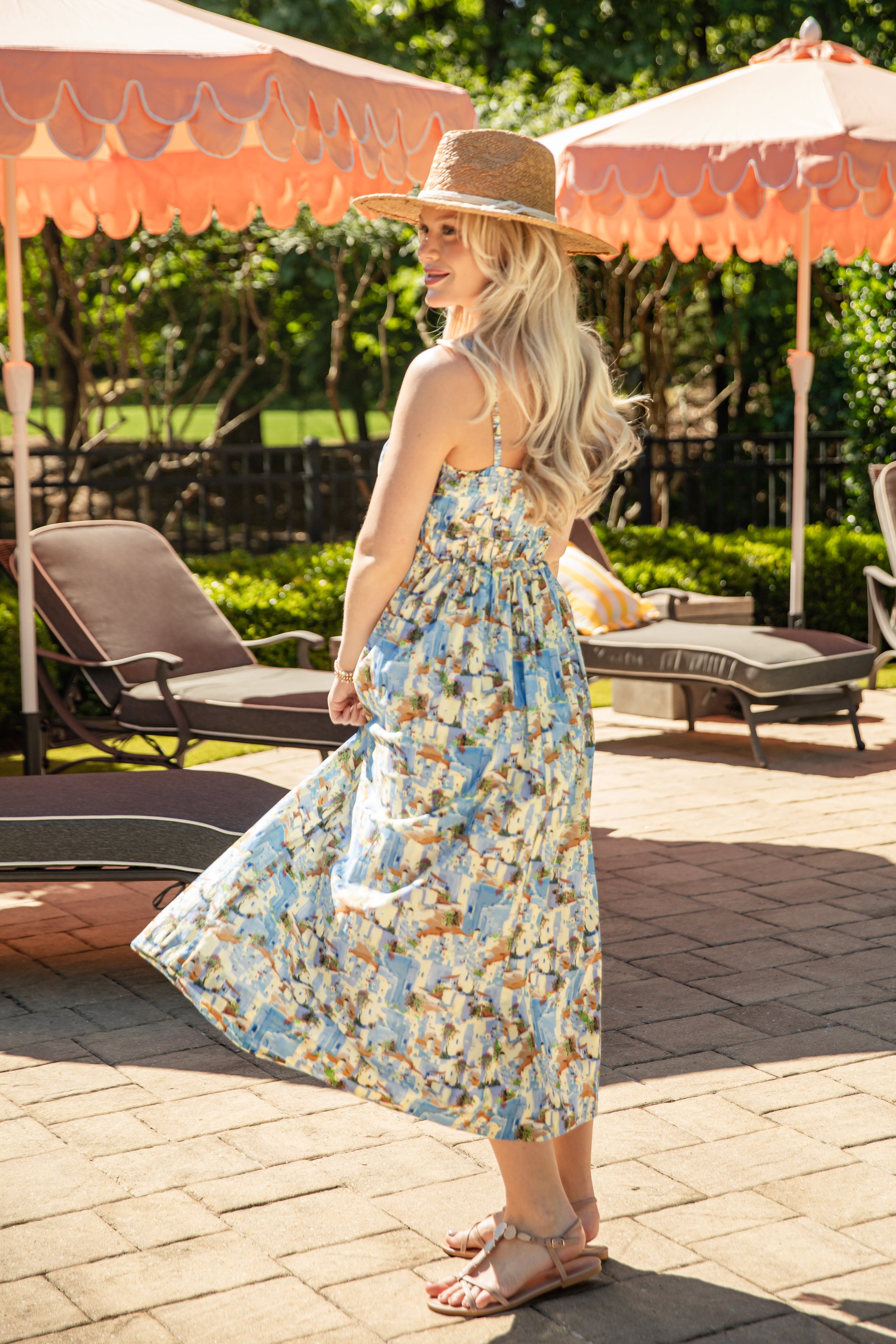 Woman in a floral dress standing under a patio umbrella with lounge chairs and greenery in the background.