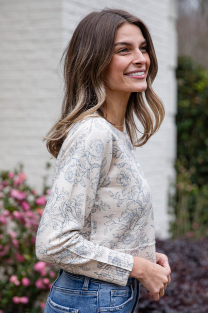 Woman wearing a floral sweater and blue jeans standing outdoors with flowers and a building in the background.