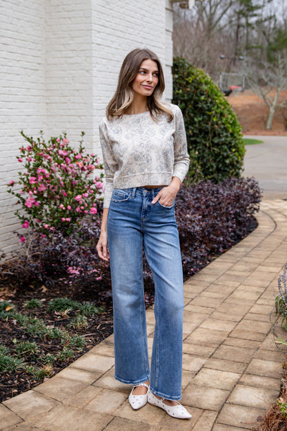 Woman wearing a light sweater and blue jeans standing outdoors near a building with plants.