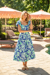 Woman in a blue floral dress standing outdoors with patio furniture and umbrellas in the background.