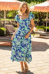 Woman in a blue floral dress standing outdoors with lounge chairs and umbrellas in the background.