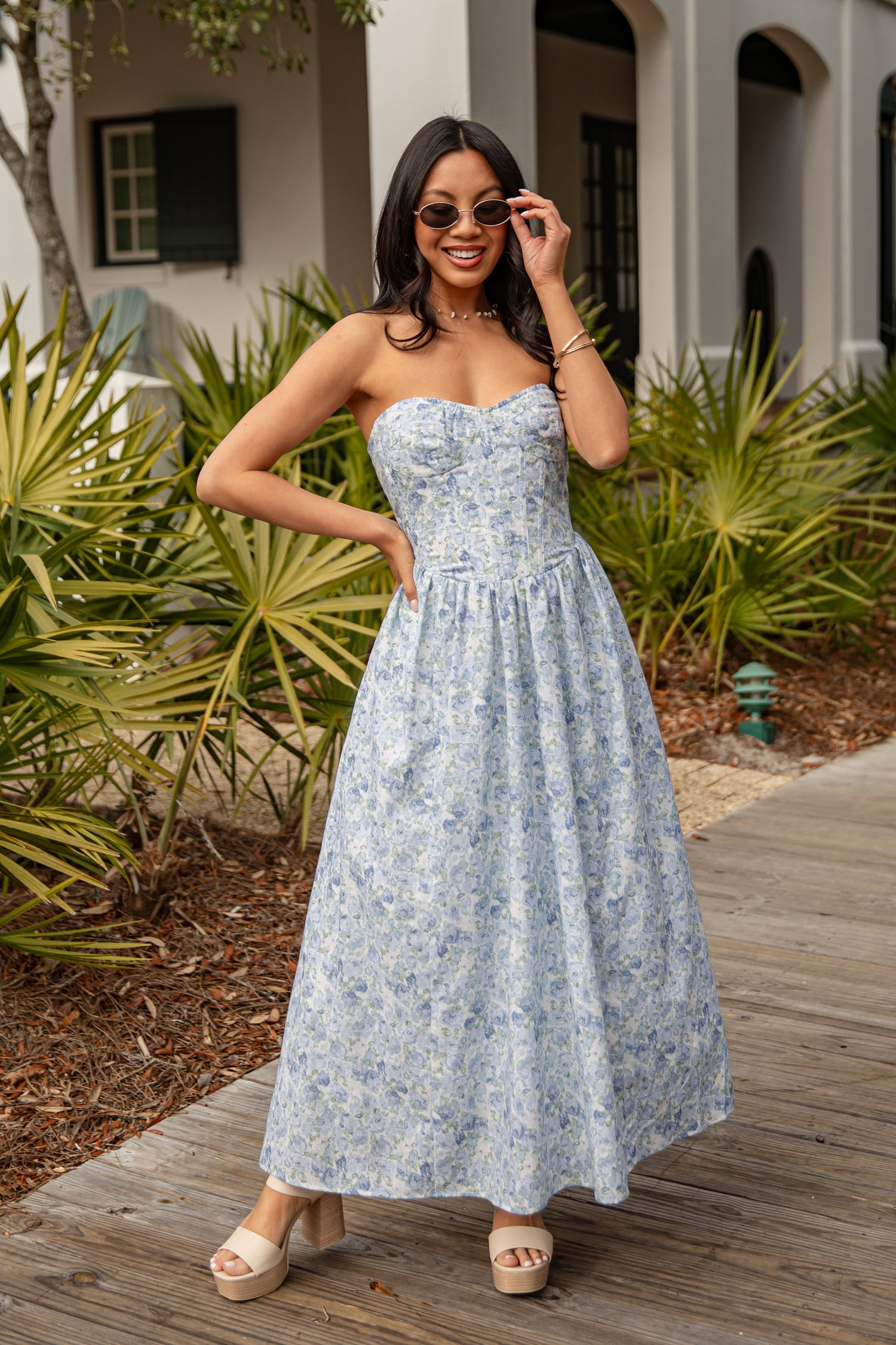 Woman in a strapless floral dress standing outdoors with plants and a building in the background