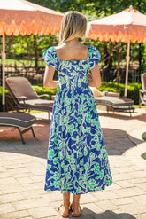 Woman in a blue floral dress standing in a sunlit outdoor setting with lounge chairs and umbrellas.