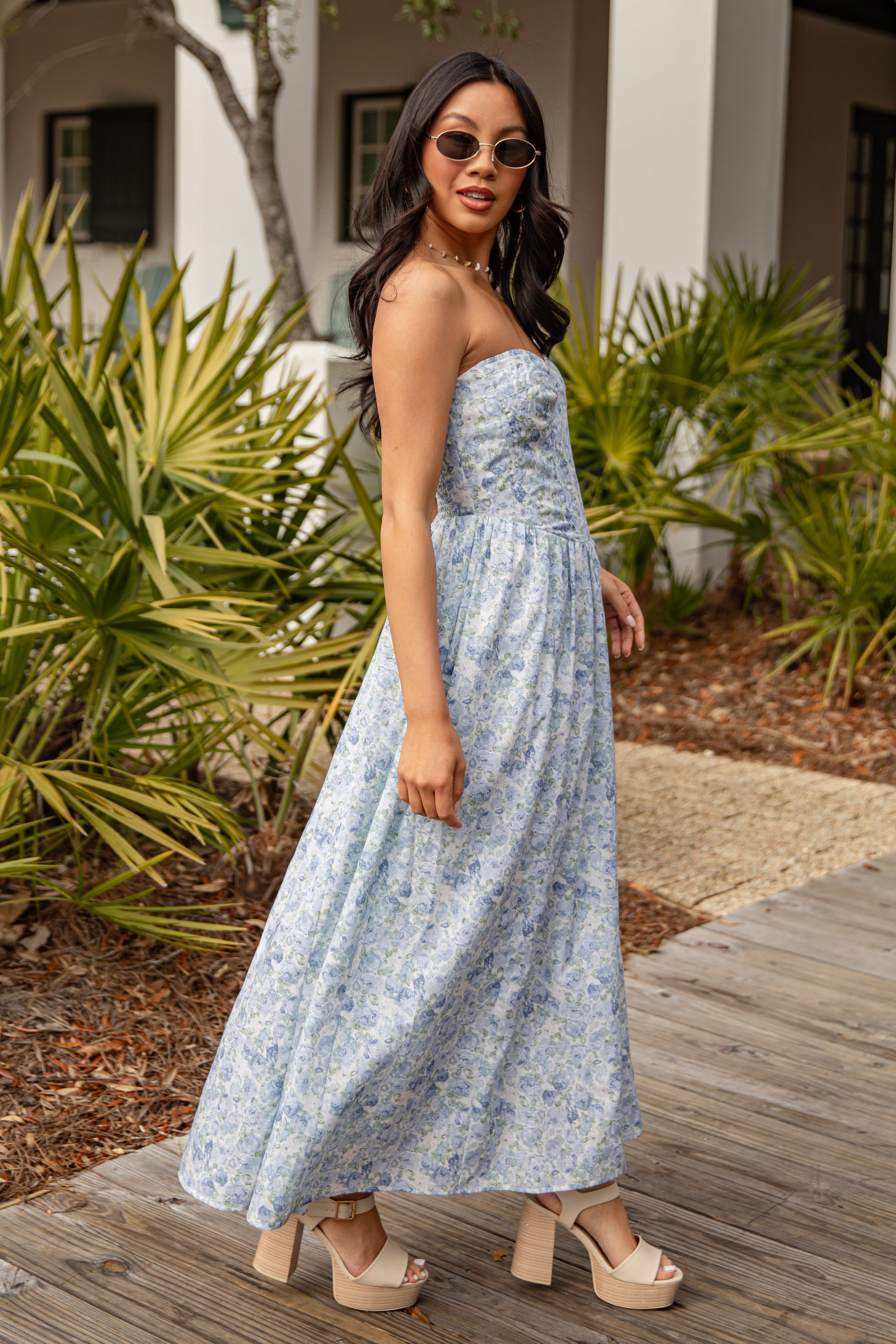 Woman in a strapless floral dress standing outdoors with plants and a building in the background