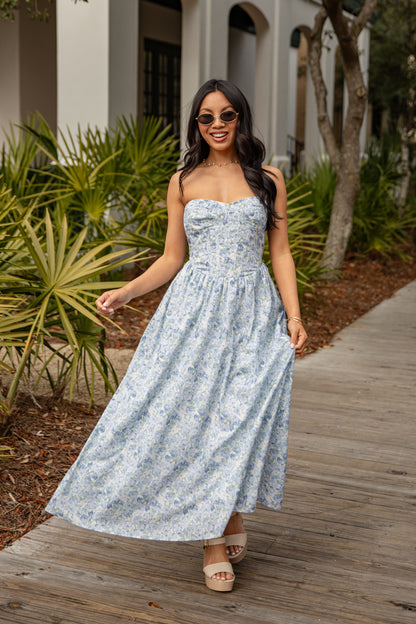 Woman in a floral dress walking on a wooden path with plants and a building in the background