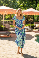 Woman in a blue floral dress standing outdoors near lounge chairs and umbrellas.