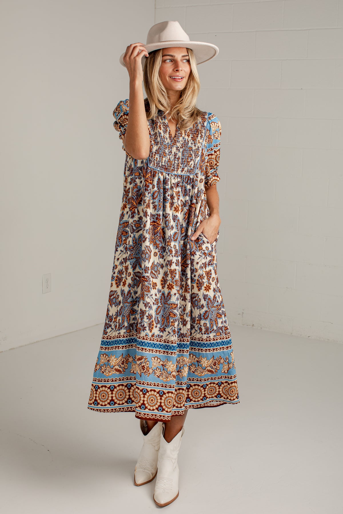 A woman modeling a bohemian-style midi dress with a brown, blue, and beige floral pattern, paired with a beige wide-brim hat and white cowboy boots, standing against a white studio wall.
