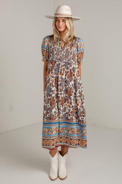 A woman modeling a bohemian-style midi dress with a multicolor floral and geometric pattern in blue, orange, brown, and cream. She is wearing a cream wide-brim hat and white cowboy boots, standing against a white brick wall.